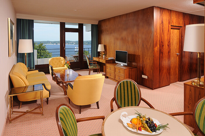 Junior Suite del Maritim Hotel Kiel, accogliente zona soggiorno, scrivania, TV e balcone con vista sull'acqua.