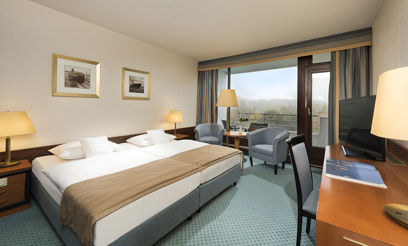 Camera classica del Maritim Hotel Kiel con ampio letto matrimoniale, area salotto, scrivania e balcone con vista sul verde.